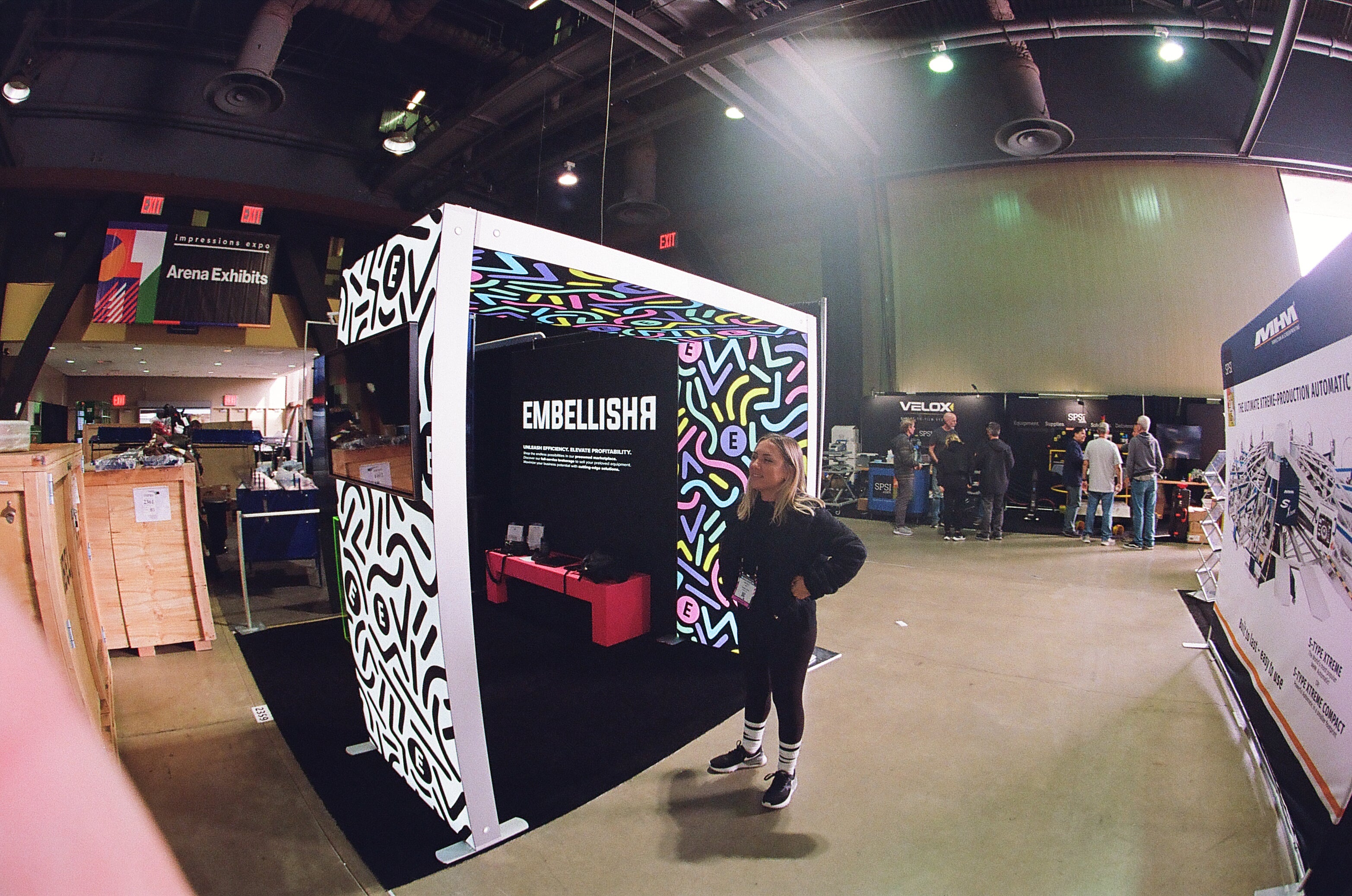 Nicole Pape standing in front of the Embellishr Tradeshow booth with colorful design and a pink bench beside the MHM booth